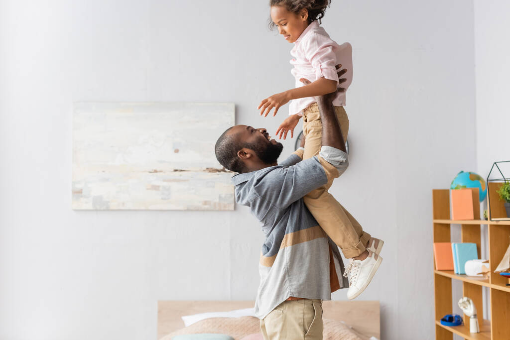 side view of young african american man raising daughter above head while having fun at home - Photo, Image