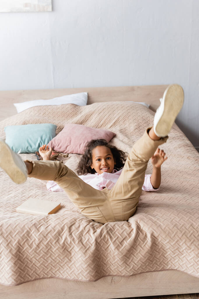 excitada chica afroamericana acostada en la cama con las piernas levantadas mientras se divierten en el dormitorio - Foto, imagen