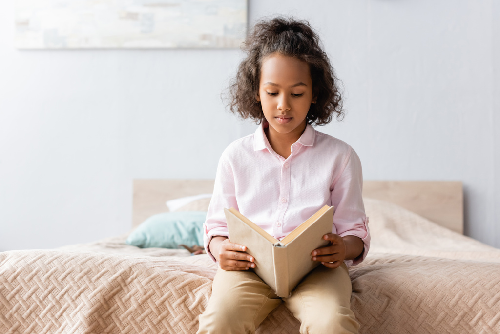 концентрована афроамериканська дівчина в білій сорочці читає книгу, сидячи на ліжку
 - Фото, зображення