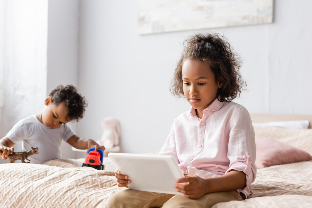 Erkek kardeşinin yanında dijital tablet kullanan konsantre Afro-Amerikan kız, yatak odasında oyuncaklarla oynuyor. - Fotoğraf, Görsel