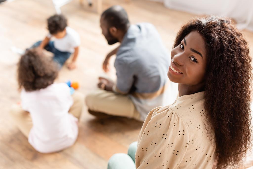 вибірковий фокус афроамериканської жінки, дивлячись на камеру біля сім'ї, що грає на підлозі
 - Фото, зображення