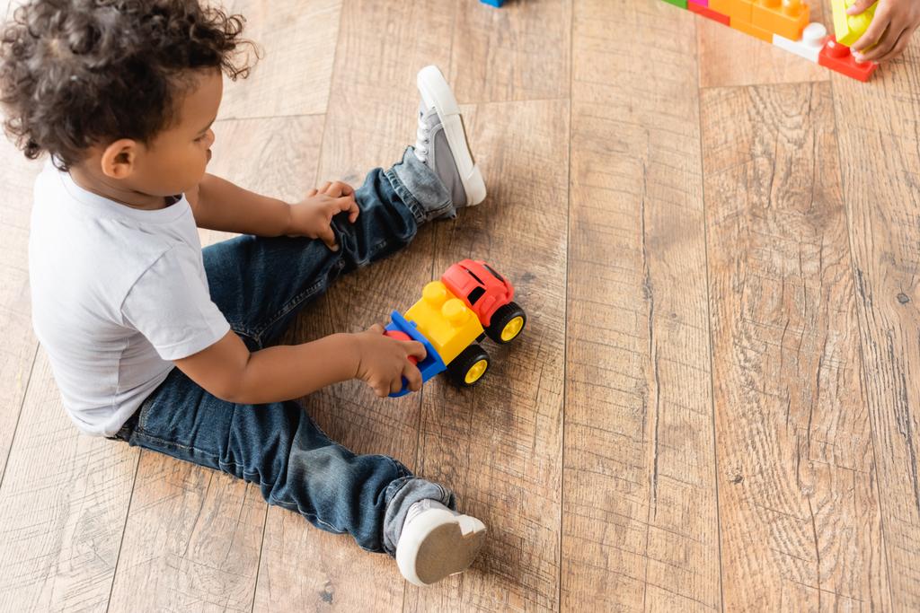 vista aérea de niño afroamericano en jeans jugando con camión de juguete en piso de madera - Foto, Imagen