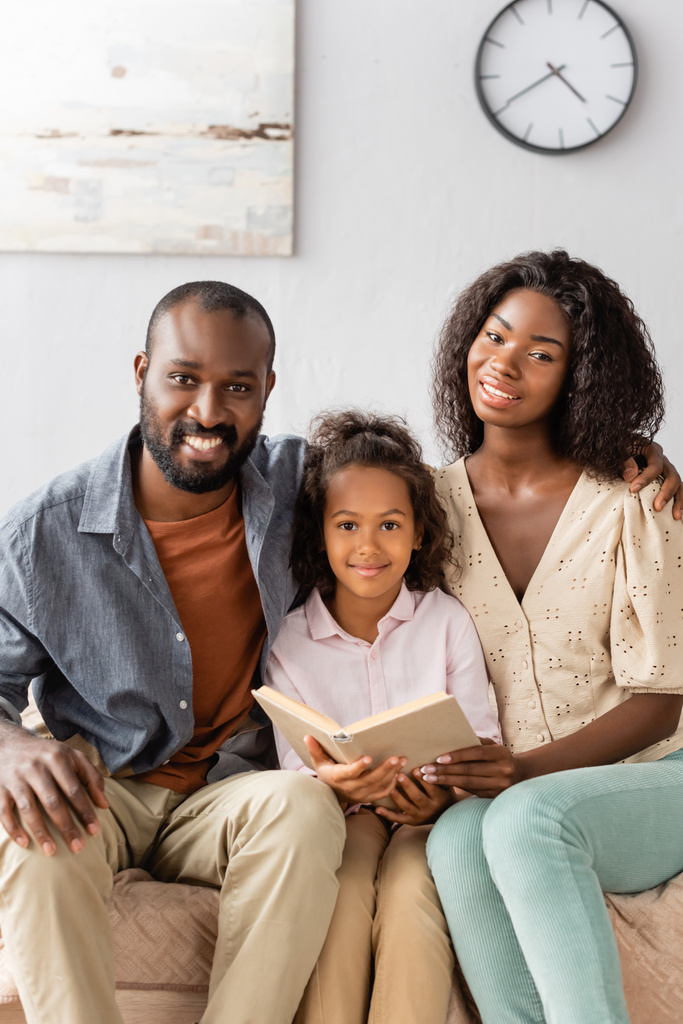 νέοι αφροαμερικανοί γονείς και παιδί με το βιβλίο κοιτάζοντας κάμερα ενώ κάθεται στο σπίτι - Φωτογραφία, εικόνα