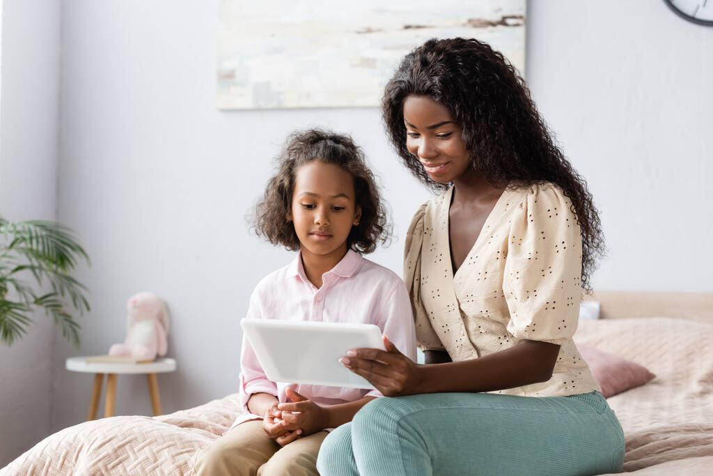 junge afrikanisch-amerikanische Frau benutzt digitales Tablet in der Nähe ihrer Tochter im Schlafzimmer - Foto, Bild