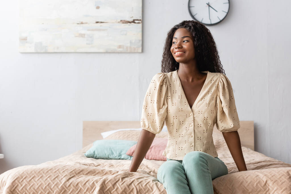 νεαρή Αφροαμερικανή γυναίκα με καθημερινά ρούχα κάθεται στο κρεβάτι και κοιτάζει αλλού - Φωτογραφία, εικόνα