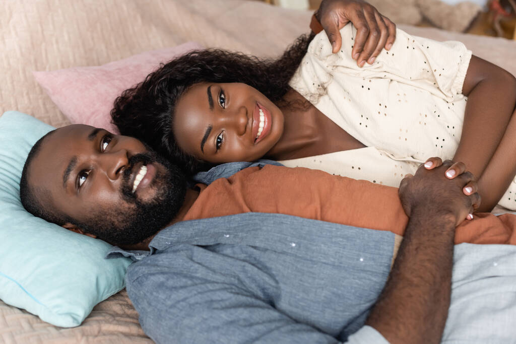high angle view of african american man embracing wife looking at camera while lying in bed - Photo, Image