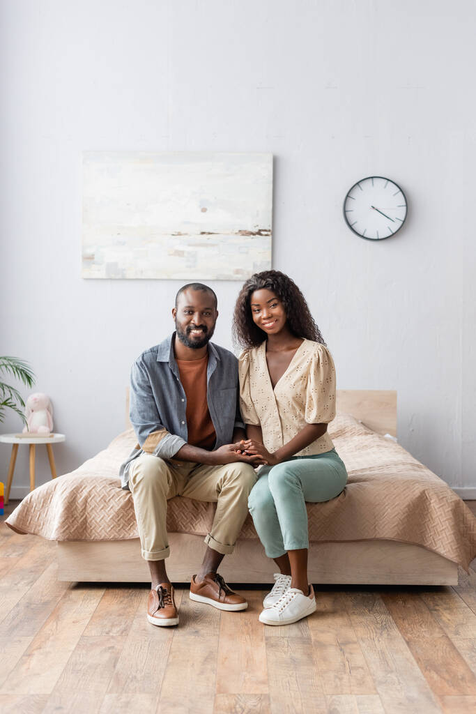 afro-amerikai férj és feleség alkalmi ruhák fogják egymás kezét, és nézi a kamera a hálószobában - Fotó, kép