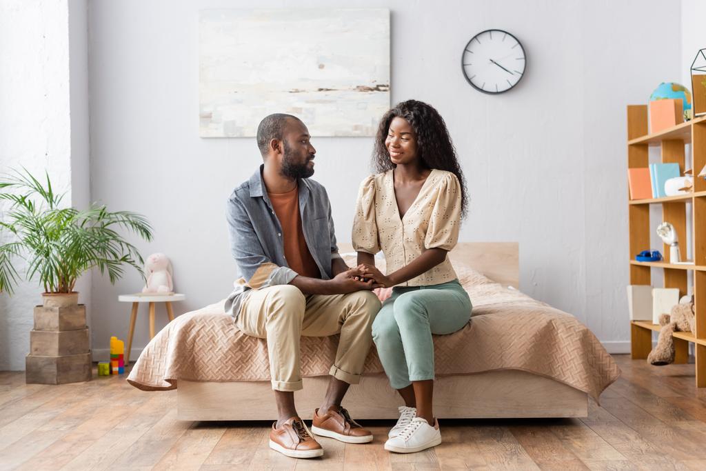 joven africano americano pareja en casual ropa cogido de la mano y mirando uno al otro en dormitorio - Foto, imagen