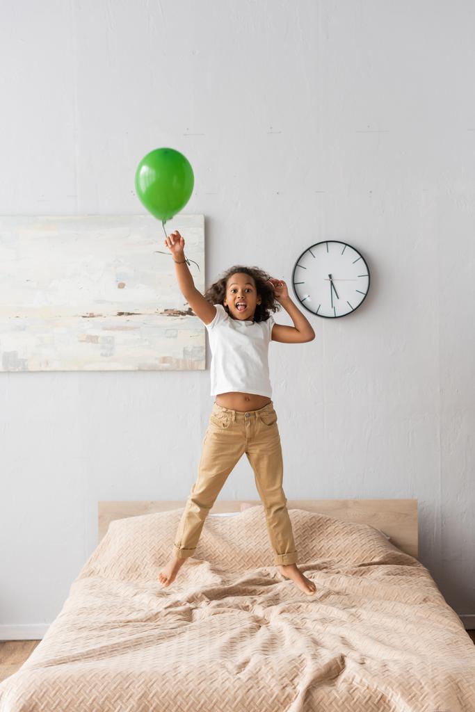 excitada chica afroamericana en ropa casual saltando en la cama con globo rojo  - Foto, imagen