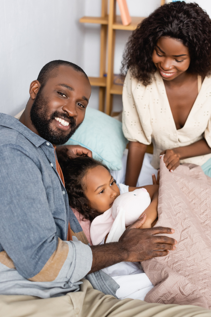 jovem afro-americano pais sentado perto criança deitado sob cobertor com brinquedo coelho - Foto, Imagem