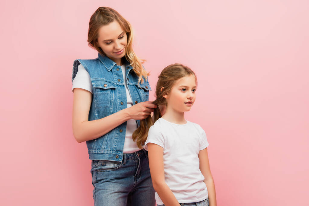 young woman in denim vest and jeans braiding hair of daughter isolated on pink - Photo, Image