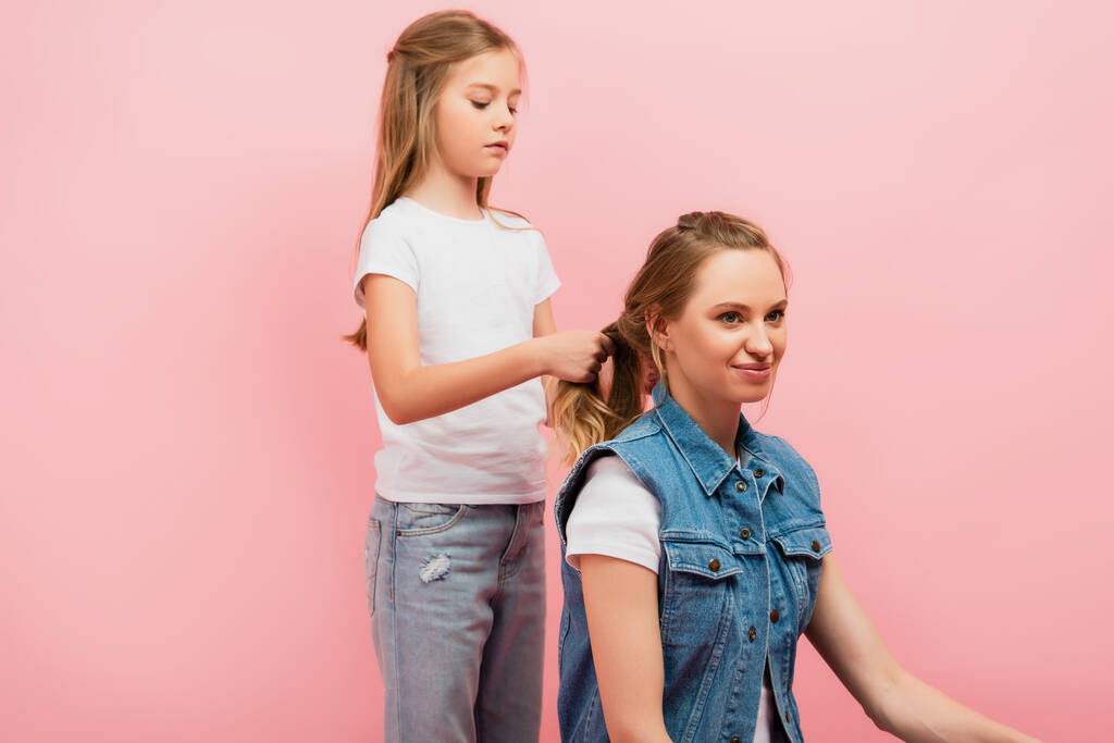 концентрированная девушка в белой футболке и джинсах плетение волос матери изолированы на розовый - Фото, изображение