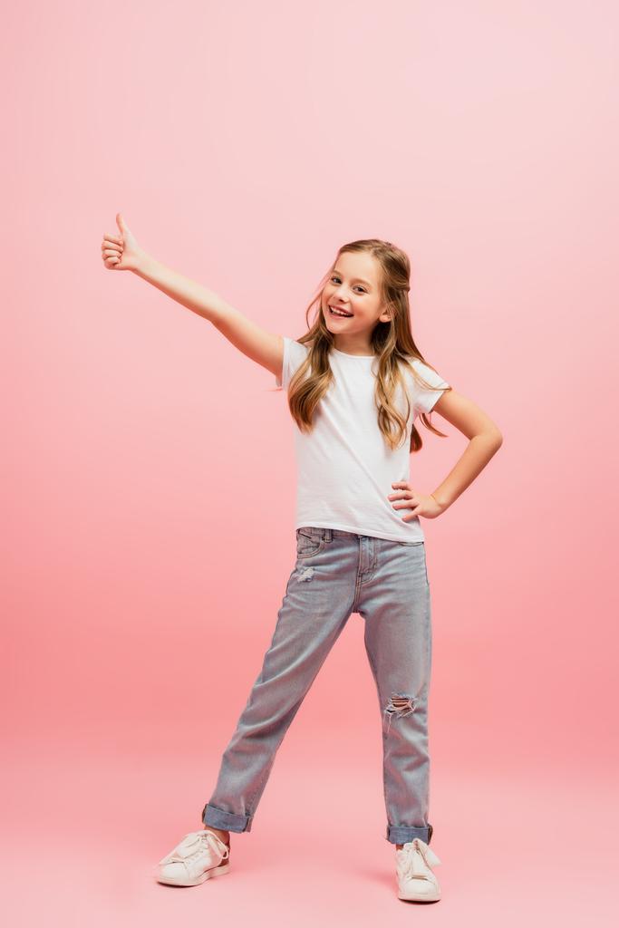 παιδί σε λευκό t-shirt με το χέρι στο ισχίο δείχνει τον αντίχειρα προς τα πάνω, ενώ κοιτάζοντας κάμερα σε ροζ - Φωτογραφία, εικόνα