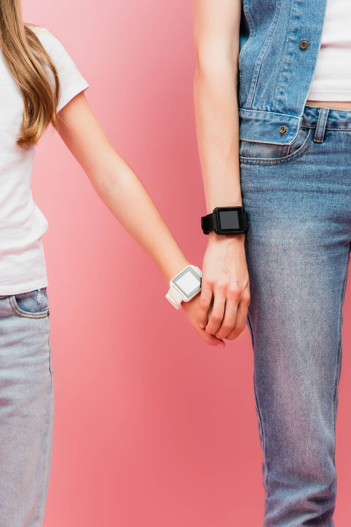 cropped view of mother and daughter in jeans and smartwatches holding hands on pink - Photo, Image