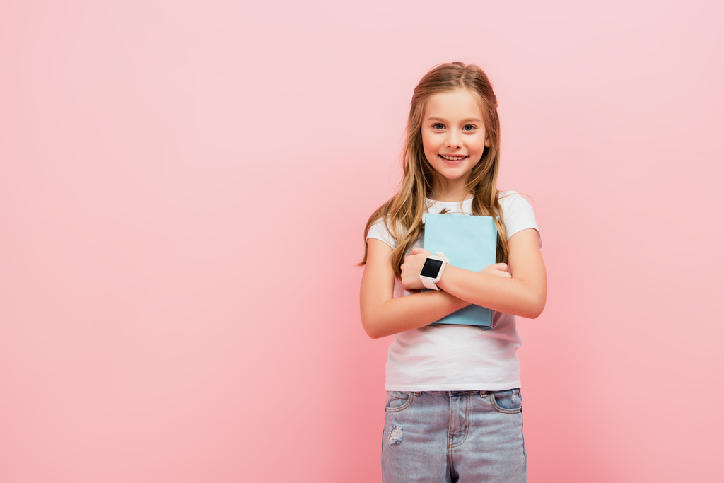 ребенок в белой футболке и смарт-часы, глядя в камеру, держа книгу изолированы на розовый - Фото, изображение