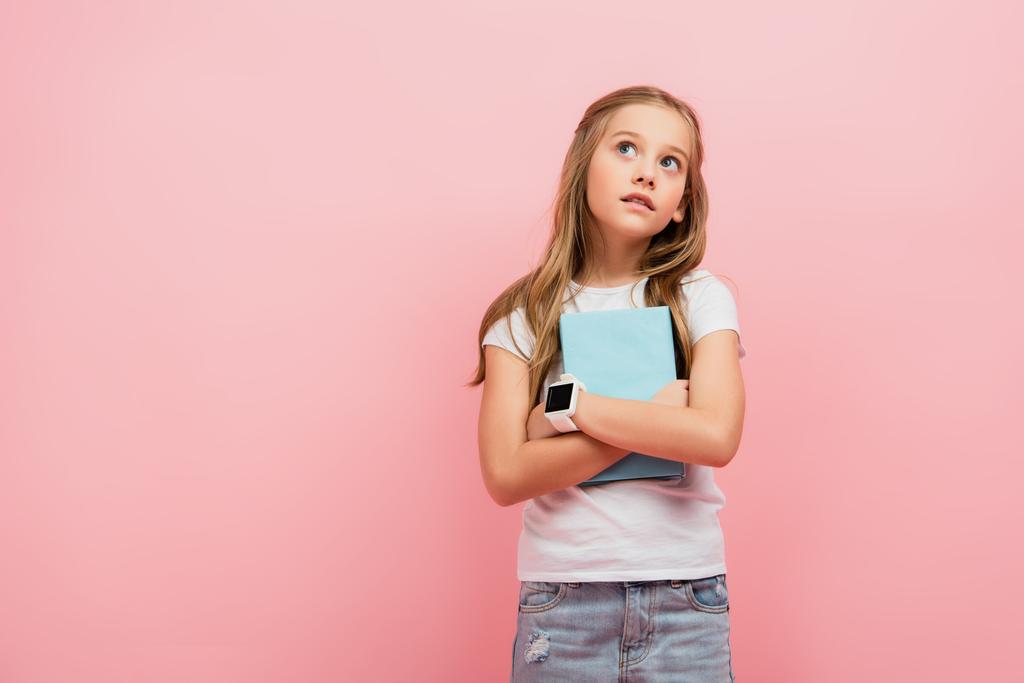 enfant réfléchi dans smartwatch regarder loin tout en tenant livre isolé sur rose - Photo, image