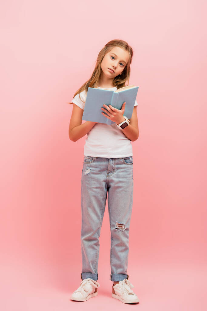 vista completa de la chica seria en smartwatch y jeans mirando a la cámara mientras sostiene el libro en rosa - Foto, imagen