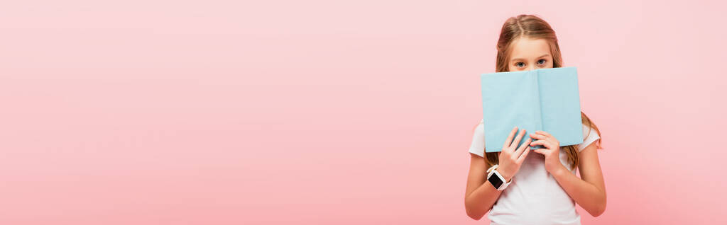 imagem horizontal da menina em smartwatch obscurecendo o rosto com livro enquanto olha para a câmera isolada em rosa - Foto, Imagem