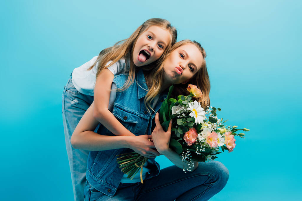 chica sobresaliendo lengua y abrazando madre puchando labios mientras sostiene ramo de flores aisladas en azul - Foto, imagen