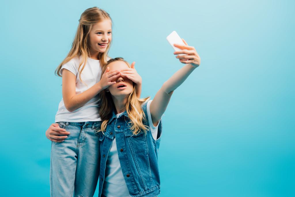 junge Frau macht Selfie auf Smartphone, während Tochter ihr Gesicht mit den Händen auf blauem Grund verdeckt - Foto, Bild
