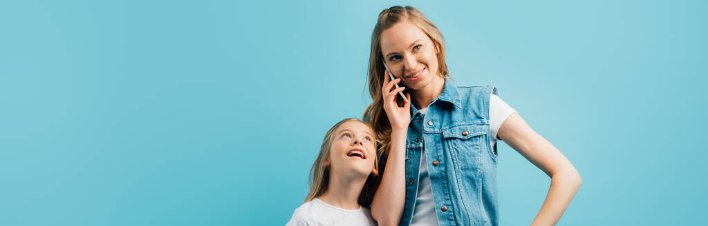 青で隔離された好奇心の強い娘の近くのスマートフォンで話すデニムベストの若い女性のパノラマショット - 写真・画像