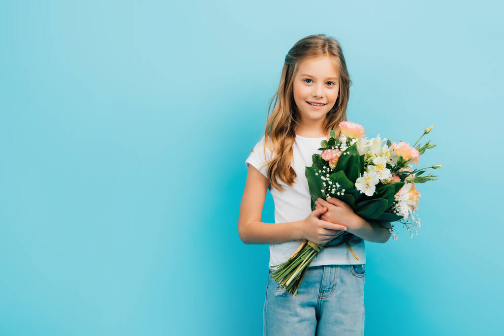 child in white t-shirt and blue jeans looking at camera while holding bouquet isolated on blue - Photo, Image