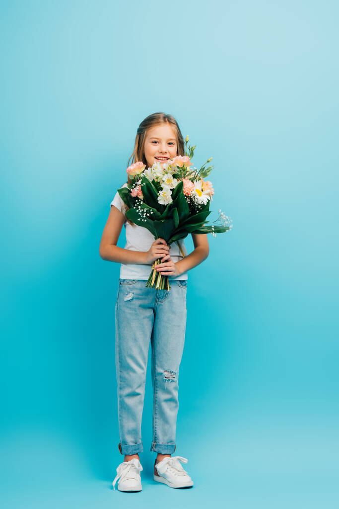 full length view of girl in jeans holding bouquet of flowers while looking at camera on blue - Photo, Image