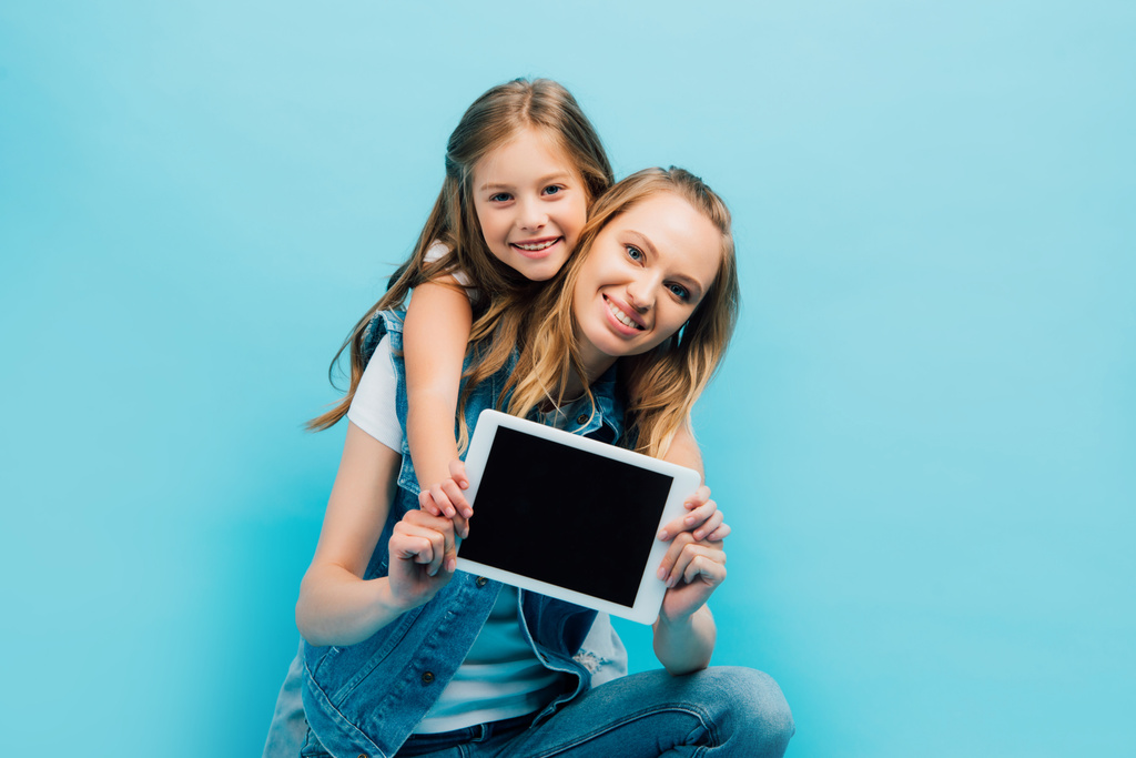 Mutter und Kind in Jeanskleidung zeigen ein digitales Tablet mit leerem Bildschirm, während sie die Kamera isoliert auf blauem Grund betrachten - Foto, Bild
