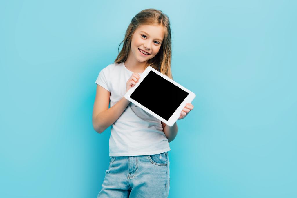 child in white t-shirt and jeans looking at camera while holding digital tablet with blank screen isolated on blue - Photo, Image