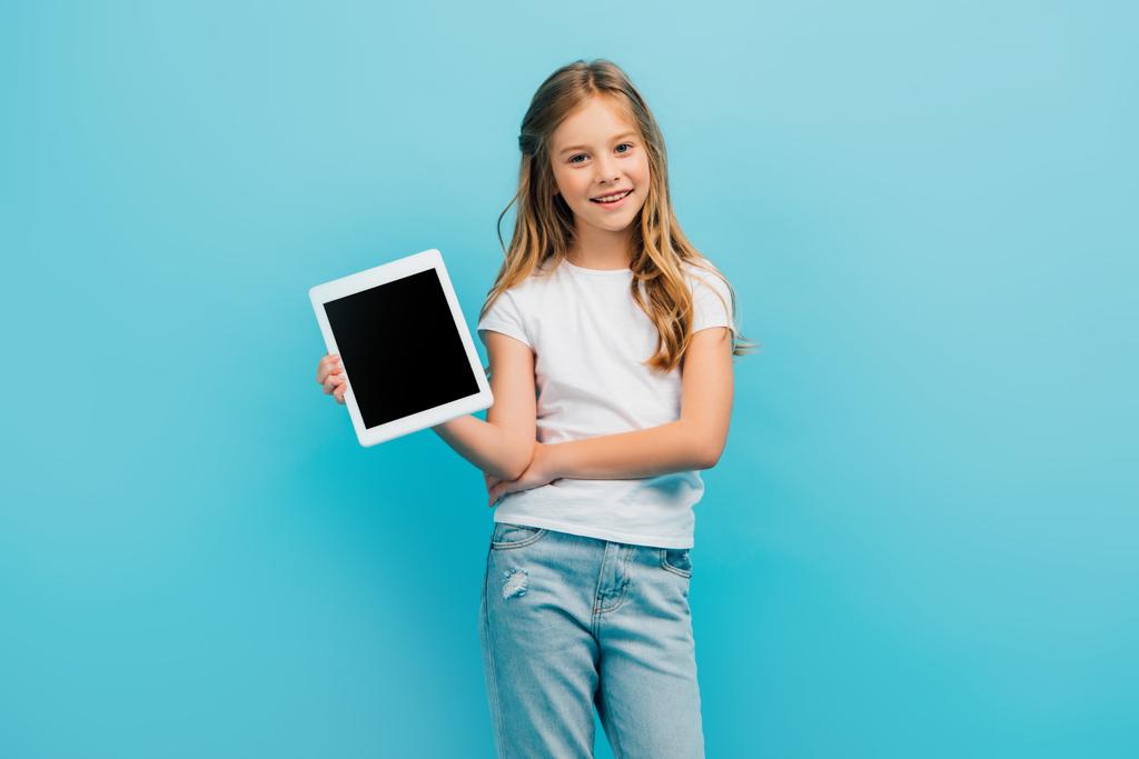 criança em camiseta branca e jeans mostrando tablet digital enquanto olha para a câmera isolada em azul - Foto, Imagem