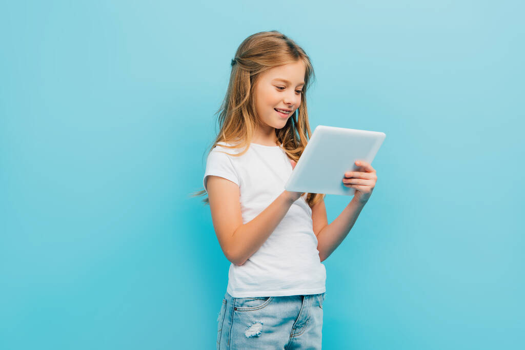 child in white t-shirt and jeans using digital tablet while standing isolated on blue - Photo, Image