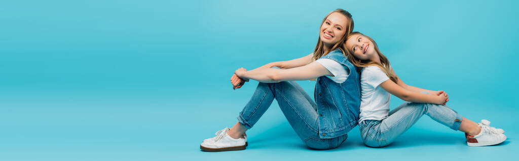 panoramic concept of young woman with daughter in denim clothes sitting back to back on blue - Photo, Image