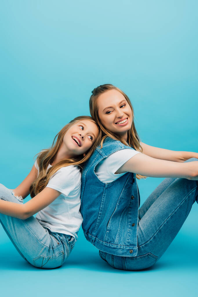 mother and daughter in denim clothes sitting on floor back to back on blue - Photo, Image