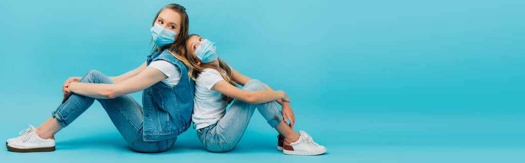 horizontal image of woman and child in denim clothes and medical masks sitting back to back on blue - Photo, Image