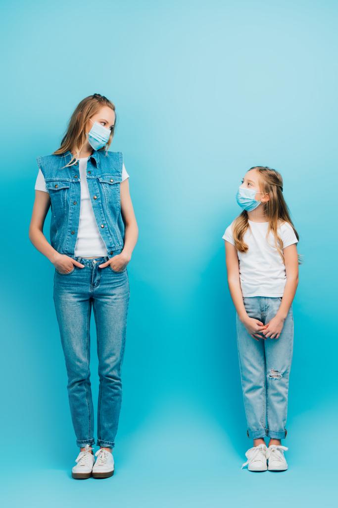 Ganzkörperansicht von Frau und Tochter in Jeanskleidung und medizinischen Masken, die einander anschauen, während sie auf blauem Grund stehen - Foto, Bild