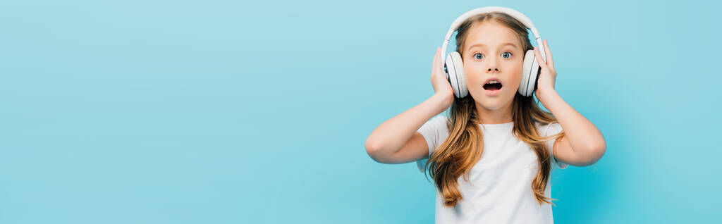 zszokowana dziewczyna w białej koszulce dotykająca bezprzewodowych słuchawek odizolowanych od niebieskiego, panoramicznego konceptu - Zdjęcie, obraz