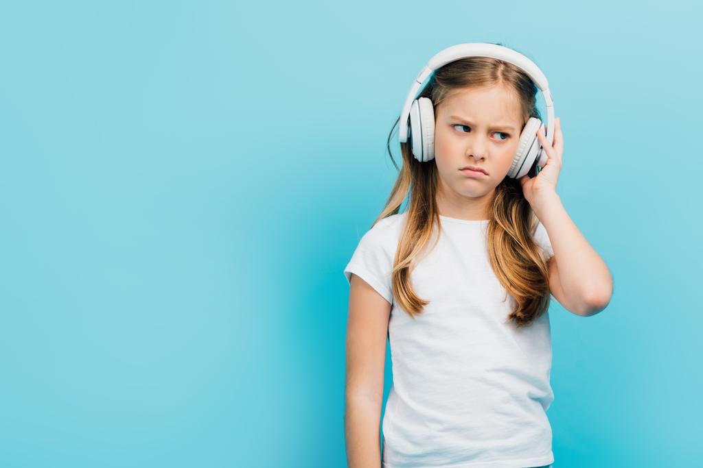 displeased girl in white t-shirt touching wireless headphones isolated on blue - Photo, Image
