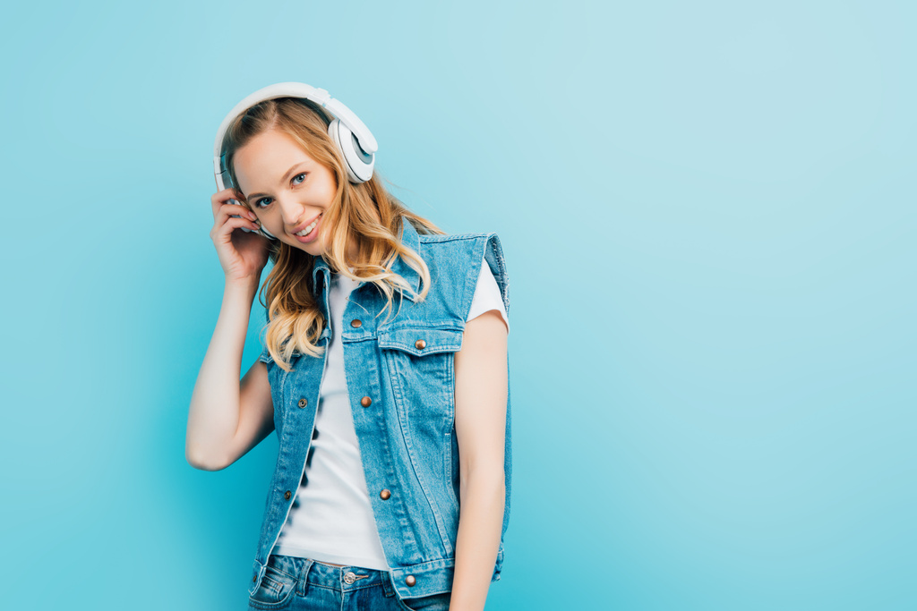 young woman in denim vest touching wireless headphones while looking at camera isolated on blue - Photo, Image