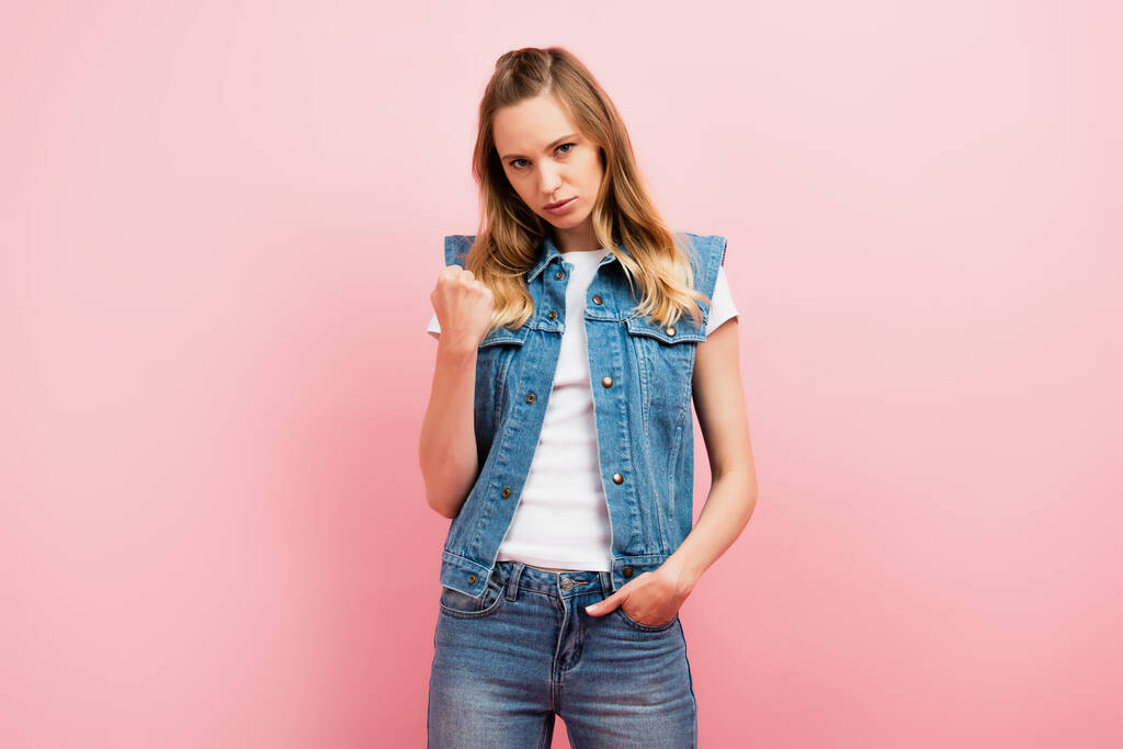 irritated woman in denim clothes showing clenched fist while standing with hand in pocket isolated on pink - Photo, Image