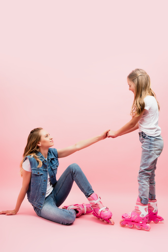 daughter holding hand of fallen mother sitting on floor in roller skates on pink - Photo, Image