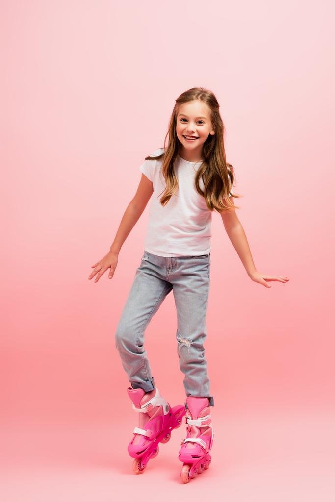 excited girl in white t-shirt, jeans and rolling skates looking at camera on pink - Photo, Image