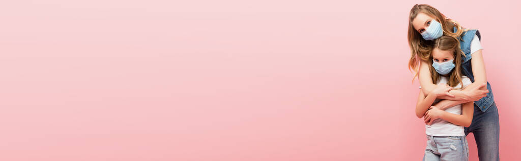 горизонтальна концепція жінки в джинсовому одязі, що обіймає доньку, носячи медичні маски разом ізольовані на рожевому
 - Фото, зображення