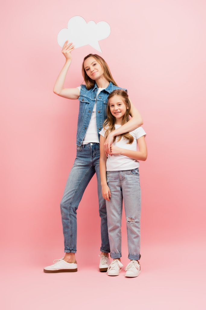vista completa de la madre en ropa de mezclilla sosteniendo la burbuja de pensamiento mientras abraza al niño en camiseta blanca y jeans en azul - Foto, Imagen