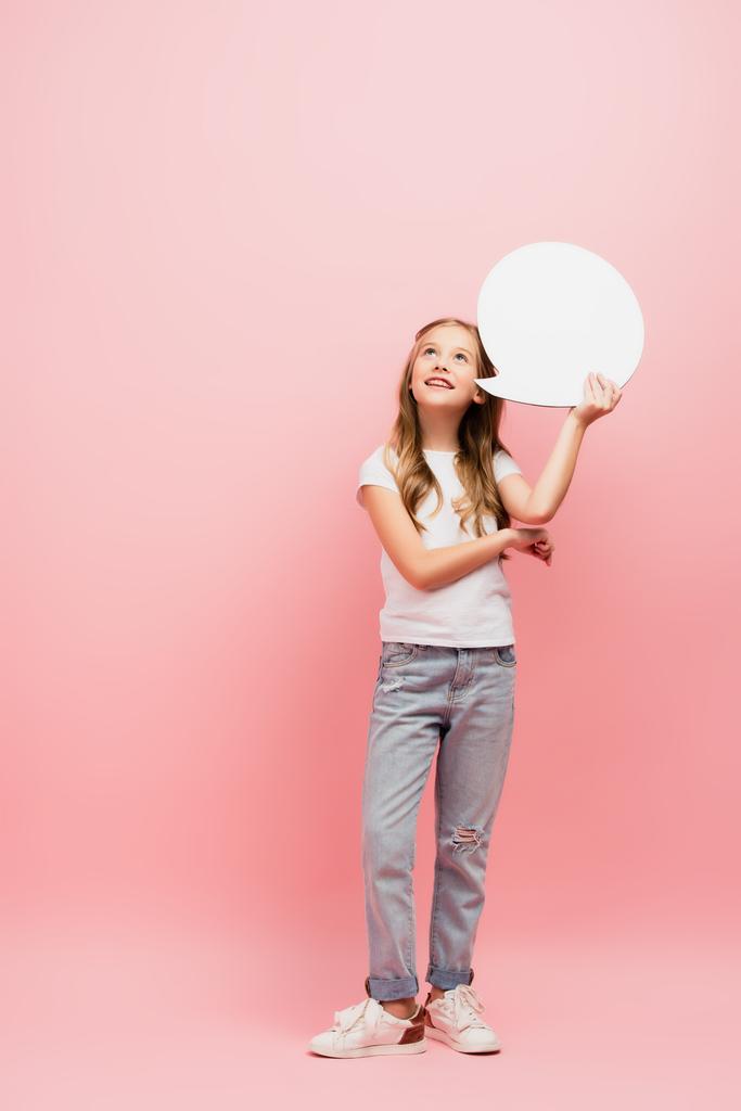 vista completa de niño en camiseta blanca y pantalones vaqueros azules mirando hacia arriba mientras sostiene la burbuja de pensamiento en rosa - Foto, imagen