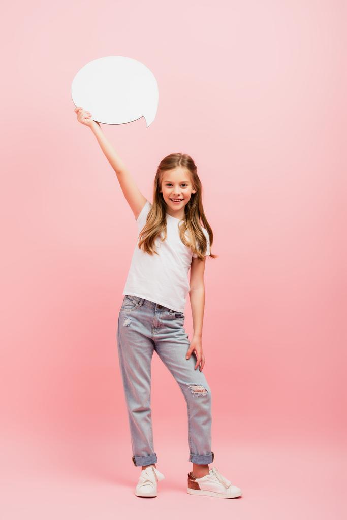 Вид в полный рост девушки в белой футболке и джинсах, держащей мыльный пузырь над головой на розовом - Фото, изображение