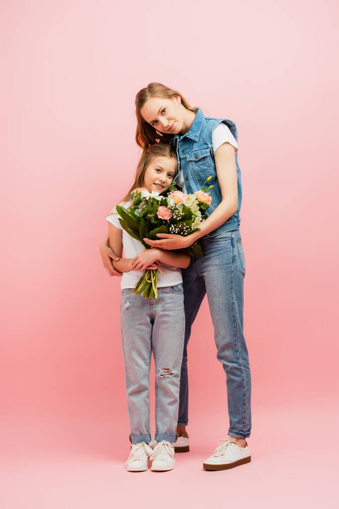 Вид в полный рост молодой женщины в джинсовой одежде, которая дарит цветы дочери и обнимает ее на розовом - Фото, изображение