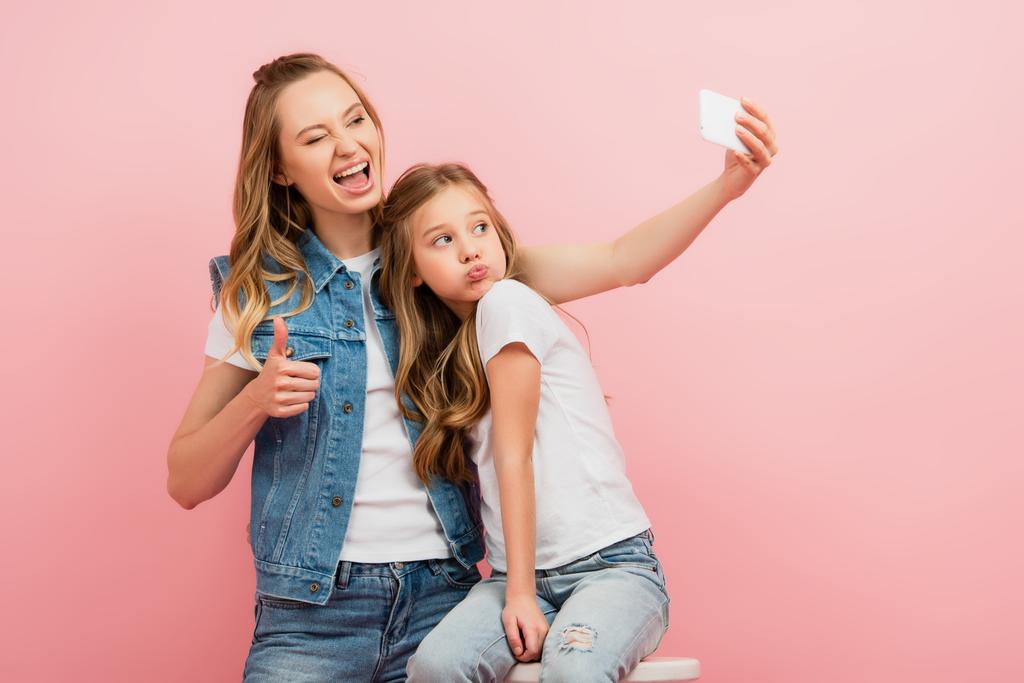 donna eccitata in denim vestiti prendendo selfie su smartphone con figlia labbra bronzanti e mostrando pollice in su isolato su rosa - Foto, immagini
