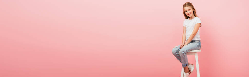 cabeçalho do site da menina em t-shirt branca e jeans azul sentado em fezes altas e olhando para a câmera isolada em rosa - Foto, Imagem