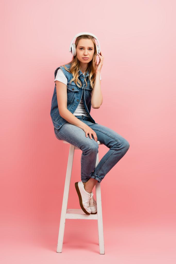 serious woman in denim clothes touching wireless headphones while sitting on high stool on pink - Photo, Image