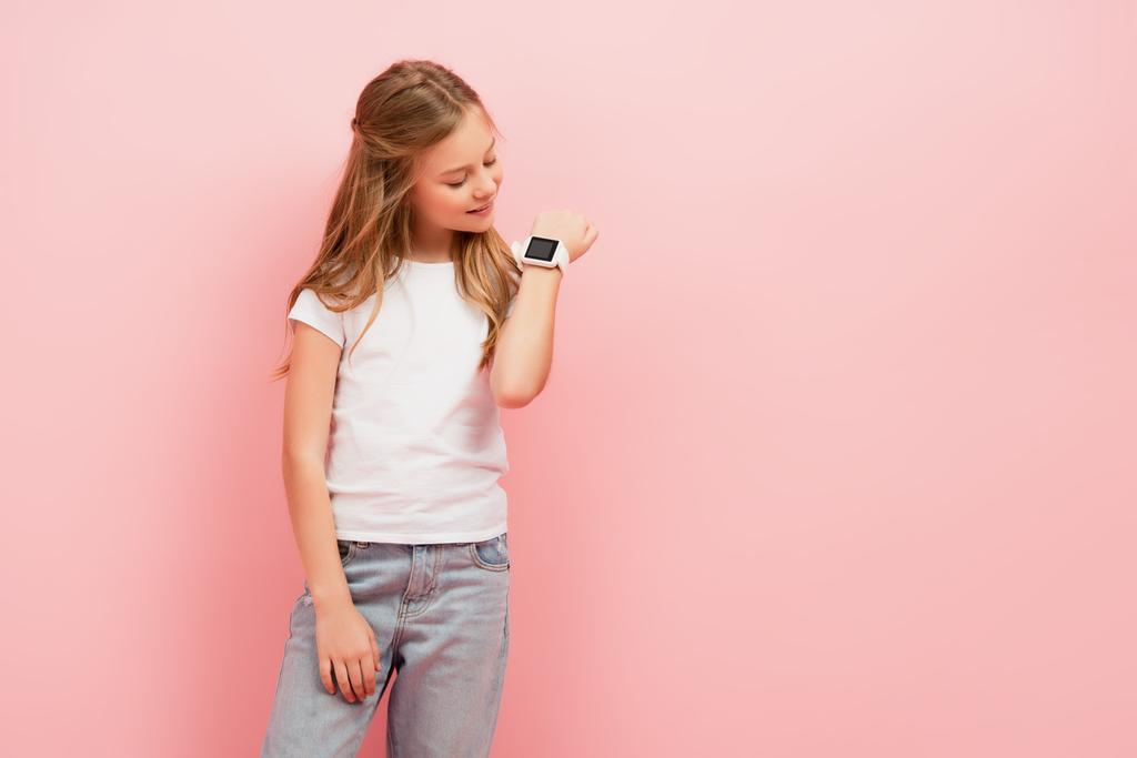 kid in wit t-shirt en jeans kijken naar smartwatch geïsoleerd op roze - Foto, afbeelding